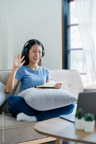 Asian young business woman is sitting on the sofa is wearing headphones video call conference with laptop computer raise hand saying hi team at home office, work from home.