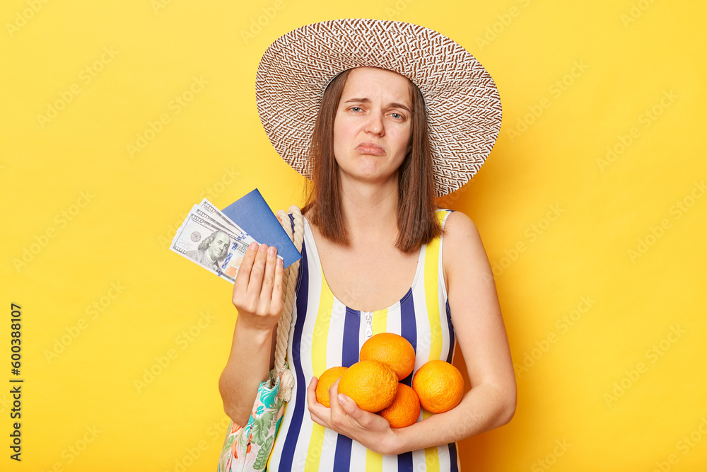 Stressed woman on resort dressed in striped swimwear and straw hat posing isolated over yellow background holding fruit and documents with money being sad her vacation canceled. Stressed woman on resort dressed in striped swimwear and straw hat posing isolated over yellow background holding fruit and documents with money being sad her vacation canceled.