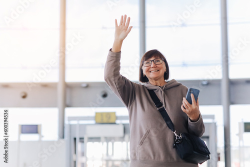 Happy smiling mature traveler woman hold smartphone and wave hand in greeting someone who meeting her at airport terminal after arrived from trip at new country at holiday vacation. Retirement travel