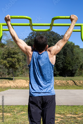 Middle-aged muscular man doing pull-ups on horizontal bar in outdoors gym. Back view of fit and athletic sportsman trainer doing workout on a sunny day. Health, exercise, fitness and wellness concept.