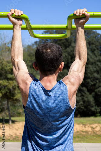 Middle-aged muscular man doing pull-ups on horizontal bar in outdoors gym. Back view of fit and athletic sportsman trainer doing workout on a sunny day. Health, exercise, fitness and wellness concept.