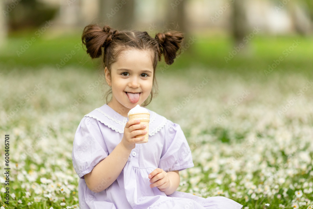 Funny happy toddler girl wearing cotton summer dress sitting on a green lawn eating vanilla and chocolate ice cream cone in a sunny garden or park. Space for text Funny happy toddler girl wearing cotton summer dress sitting on a green lawn eating vanilla and chocolate ice cream cone in a sunny garden or park. Space for text