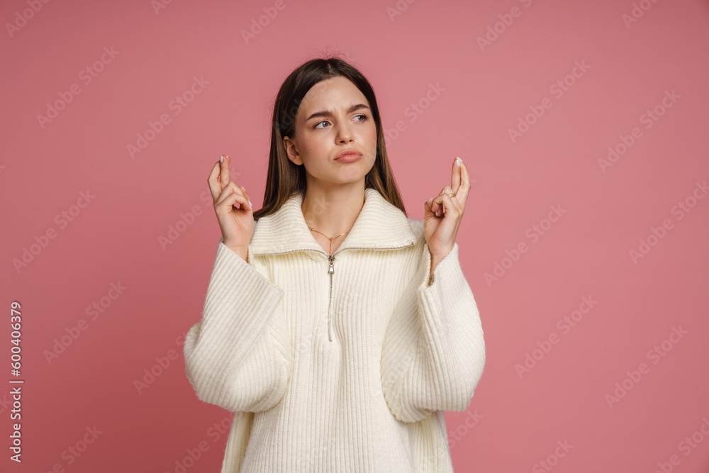 Pensive woman holding fingers crossed for good luck and looking aside isolated over pink background