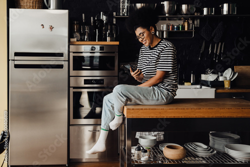 Cheerful woman using smartphone while sitting on table in kitchen