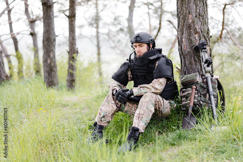 A man in a military uniform and a bulletproof vest sits in the forest near a metal detector and a military backpack. A man pauses in the work of demining the territory