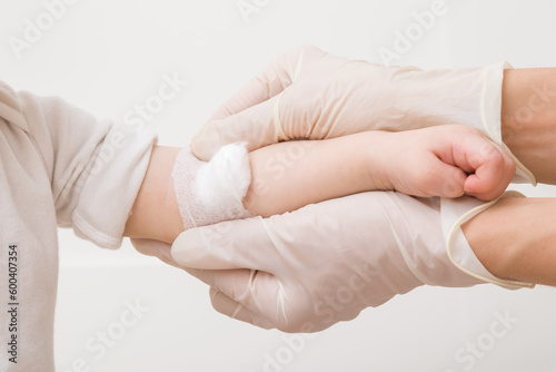 Doctor hands in white rubber protective gloves putting bandage with cotton wool on baby arm vein after blood test or injection of vaccine. Children healthcare. Closeup. Light gray background. © fotoduets