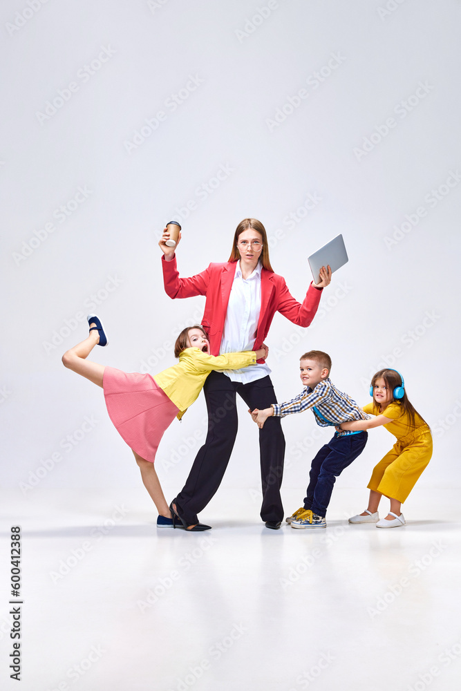 Combination of work and parenting. Portrait business woman, mother and her little kids disturbing, playing around against grey studio background. Concept of family, motherhood, childhood, lifestyle