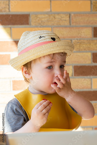Selective focus vertical view of fair toddler boy sitting in high chair on terrace stuffing his mouth with red crackers