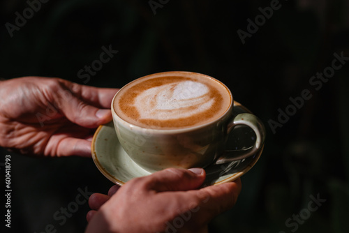 Men's hands hold a mug of cappuccino on a dark contrasting background. The barista made delicious coffee with milk. Stylish photo of ready-made coffee in the sun
