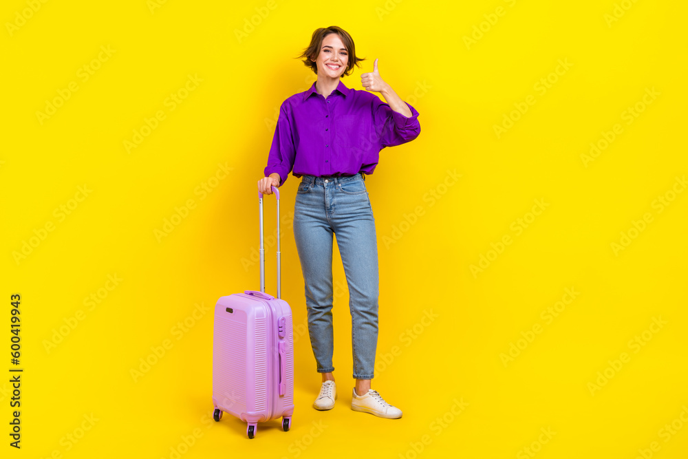 Full length photo of cute confident woman dressed purple shirt showing thumb up holding luggage isolated yellow color background Full length photo of cute confident woman dressed purple shirt showing thumb up holding luggage isolated yellow color background