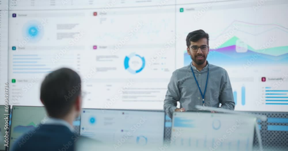 Male Indian Computer Scientist With Tablet Giving Presentation to Diverse Team Of Data Analysts In Front Of Big Digital Screen In Monitoring Room. Colleagues Listening To Training On Automation.