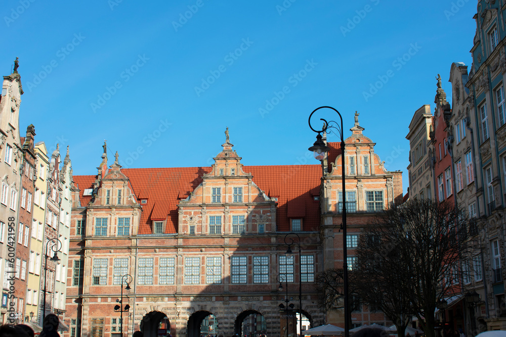 Fototapeta premium View of an ancient building with a red roof, windows and arches against a blue sky. Street lanterns and old buildings around. Central square. Old architecture. Cityscape. Poland, Gdansk, April 2023