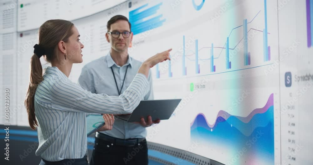 Caucasian Male And Female Data Scientists With Laptop And Tablet Standing Next To Big Digital Screen With Graphs And Charts In Monitoring Office. Colleagues Discussing Business Opportunities, Risks.