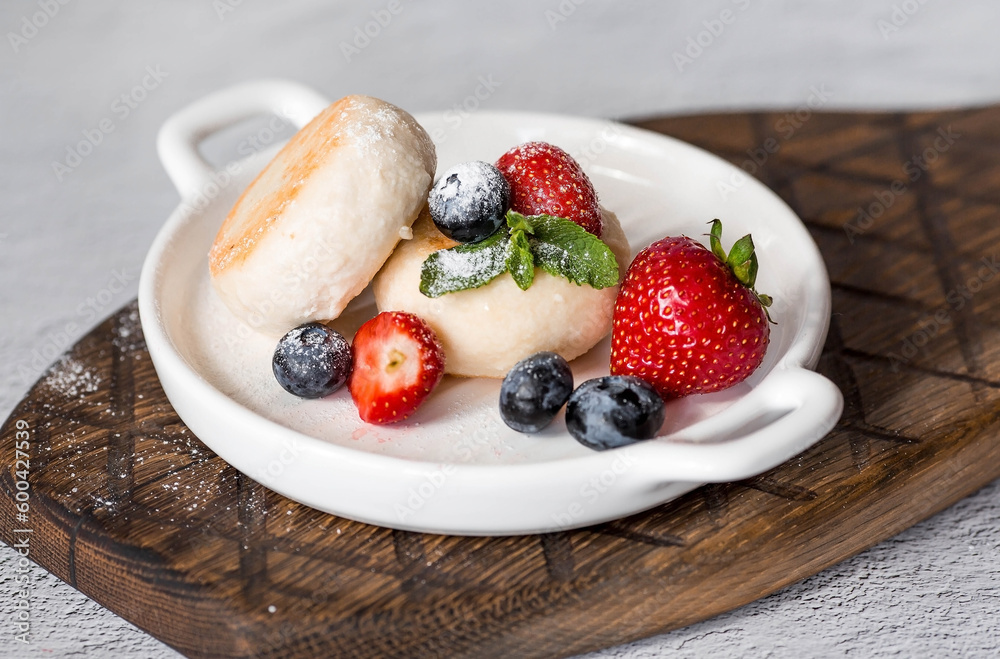 Cottage cheese pancakes, Russian cheesecakes with fresh blueberries, strawberries and mint on a stylish wooden board on a gray concrete table. Natural products. Healthy and delicious breakfast.