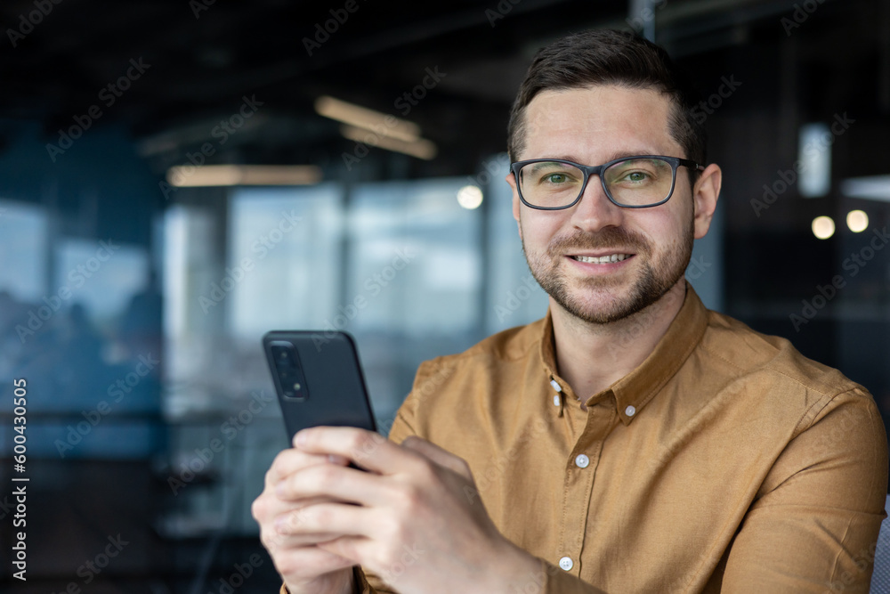 Closeup portrait of a programmer with a phone, a man in a shirt at the workplace smiles and looks at the camera, a businessman holds a phone, uses an online application, makes a call, and browses Closeup portrait of a programmer with a phone, a man in a shirt at the workplace smiles and looks at the camera, a businessman holds a phone, uses an online application, makes a call, and browses