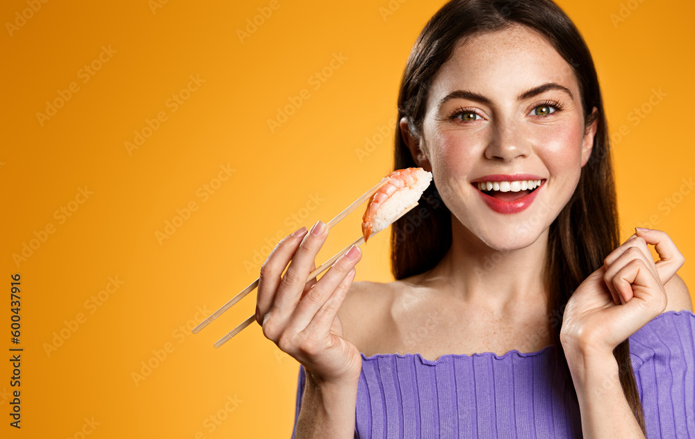 Obraz premium Smiling young woman eats japanese asian food, asian takeaway restaurant delivery, holds sushi with chopsticks, hungry girl eats sashimi, isolated on orange background
