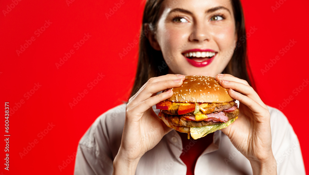 Happy young woman with big burger, bites hamburger with pleased smile, hungry face, stands isolated on red background