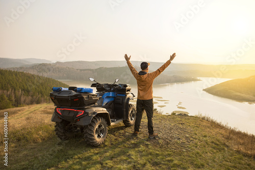 Happy man traveler raised hands at sunset mountains. Travel lifestyle. Man rider standing near ATV car. Moto tourism concept.