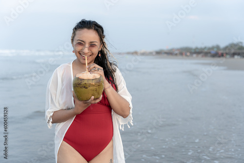 A young Hispanic woman is drinking with a coconut on the sea edge at the beach photo