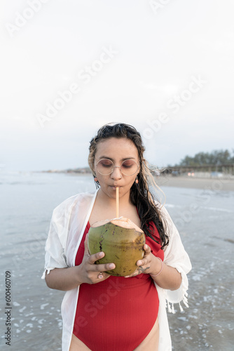 A young Hispanic woman is drinking with a coconut on the sea edge at the beach photo