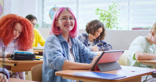 Smiling pink haired girl in glasses sitting at desk with tablet in classroom and looking at camera. Portrait of positive female student studying with male and female classmates on background.