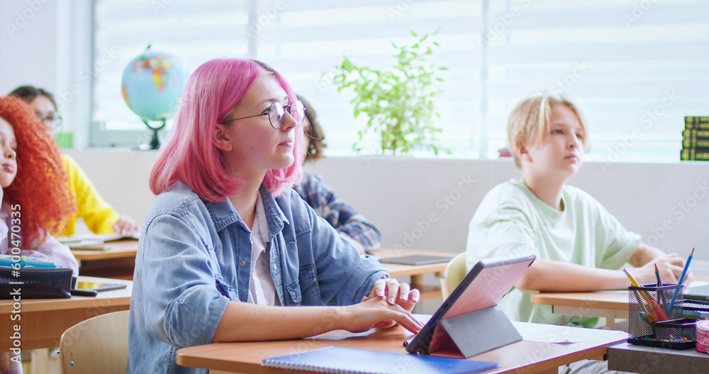 Obraz premium Portrait of happy teenage Caucasian girl in glasses and with pink hair sitting at desk with tablet device and smiling to camera. Cheerful stylish female student using computer at school. Lesson.