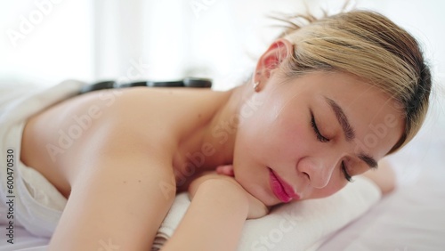 Close up face of beautiful Asian woman in white towel lying on massage bed closed eyes having hot stones on back. Wellness and body relaxation therapy 