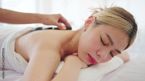 Beautiful Asian woman lying on spa bed with closed eyes and relaxed while masseuse putting hot stones on back. Spa back massage and wellness therapy