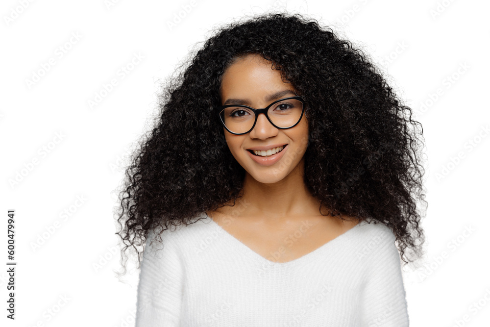 Gorgeous lovely curly woman with Afro hairstyle, feels glad, smiles gently at camera, wears optical glasses and white sweater, isolated on grey background. Happy emotions and feelings concept Gorgeous lovely curly woman with Afro hairstyle, feels glad, smiles gently at camera, wears optical glasses and white sweater, isolated on grey background. Happy emotions and feelings concept