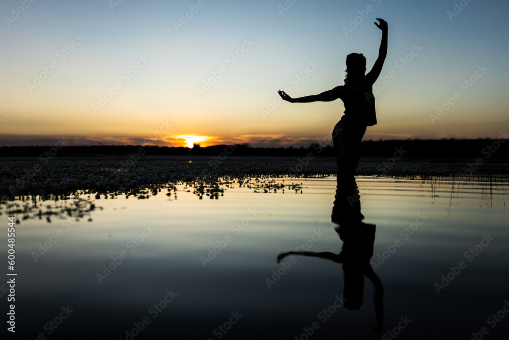 woman in the lake making figures and dancing with her body in the water, at sunset, with cold and warm colors in the sky, reflections in the water.