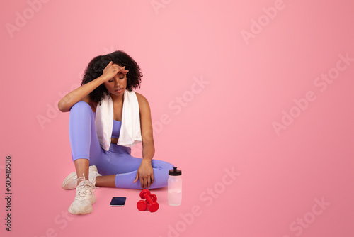 Sad tired millennial african american lady in sportswear and towel with dumbbells, bottle of water