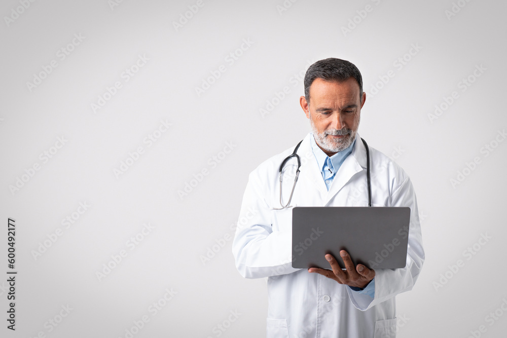 Serious confident european senior man doctor in white coat with stethoscope typing on laptop Serious confident european senior man doctor in white coat with stethoscope typing on laptop