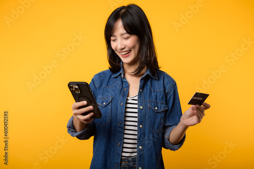 Portrait beautiful young asian woman happy smile dressed in denim jacket showing smartphone and credit card isolate on yellow studio background. Shopping online smartphone application concept