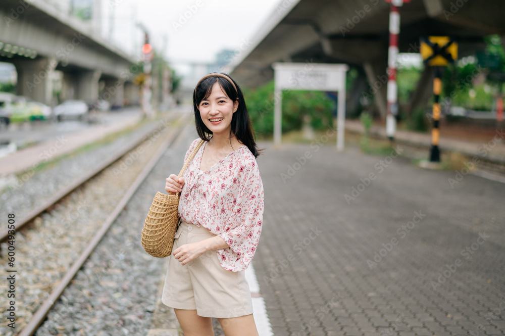Beautiful Asian young woman traveler with weaving basket standing beside railway train station in Bangkok. Journey trip lifestyle, world travel explorer or Asia summer tourism concept.
