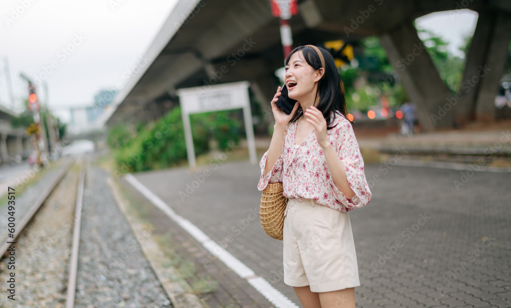 Asian young woman traveler with weaving basket using a mobile phone beside railway train station in Bangkok. Journey trip lifestyle, world travel explorer or Asia summer tourism concept. Asian young woman traveler with weaving basket using a mobile phone beside railway train station in Bangkok. Journey trip lifestyle, world travel explorer or Asia summer tourism concept.