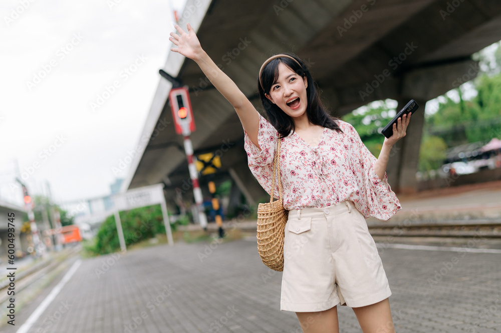 Asian young woman traveler with weaving basket using a mobile phone beside railway train station in Bangkok. Journey trip lifestyle, world travel explorer or Asia summer tourism concept.