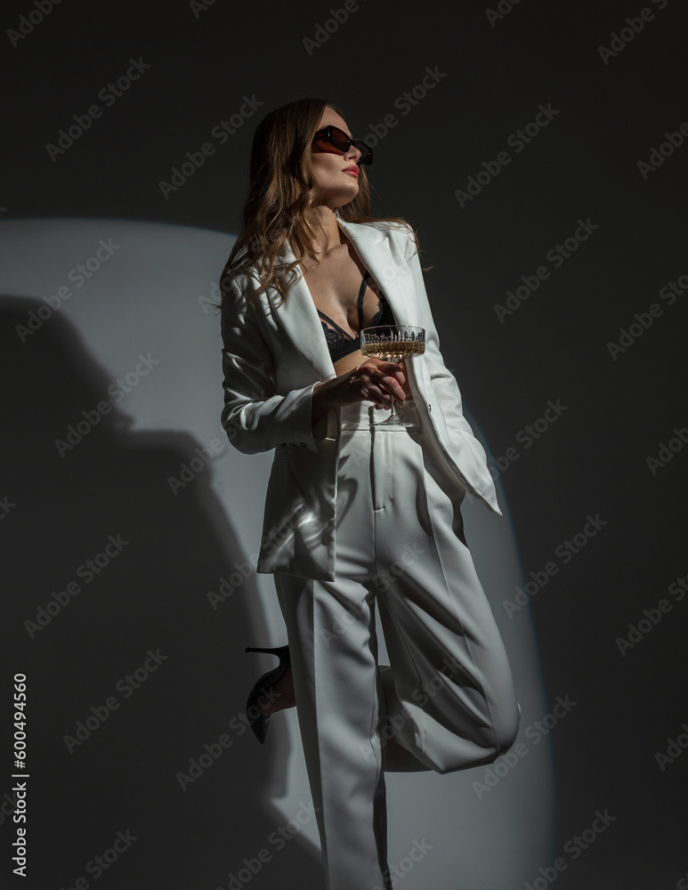 Fashion beautiful chic woman with cool sunglasses in a stylish white suit with a blazer and a bra with a glass of champagne on a dark background with a round light at a party. Lady celebrating