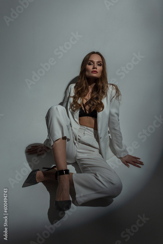 Beautiful elegant stylish chic woman model in a white fashion suit with a bra and heels sits on the floor in a studio with a hard glamour light