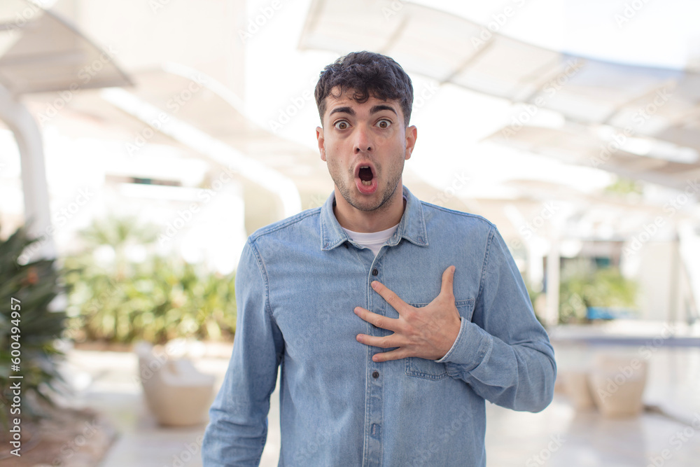 Naklejka premium young handsome man feeling shocked, astonished and surprised, with hand on chest and open mouth, saying who, me?