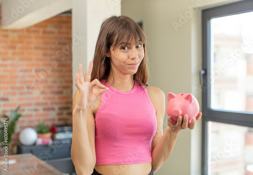 young pretty woman feeling happy, showing approval with okay gesture piggy bank concept