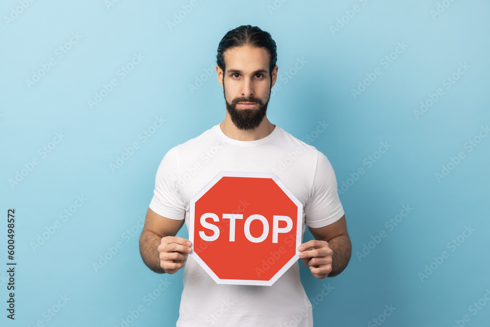 Portrait of serious man with beard wearing white T-shirt holding red Stop sign, looking at camera with negative aggressive expression, showing ban. Indoor studio shot isolated on blue background. Portrait of serious man with beard wearing white T-shirt holding red Stop sign, looking at camera with negative aggressive expression, showing ban. Indoor studio shot isolated on blue background.