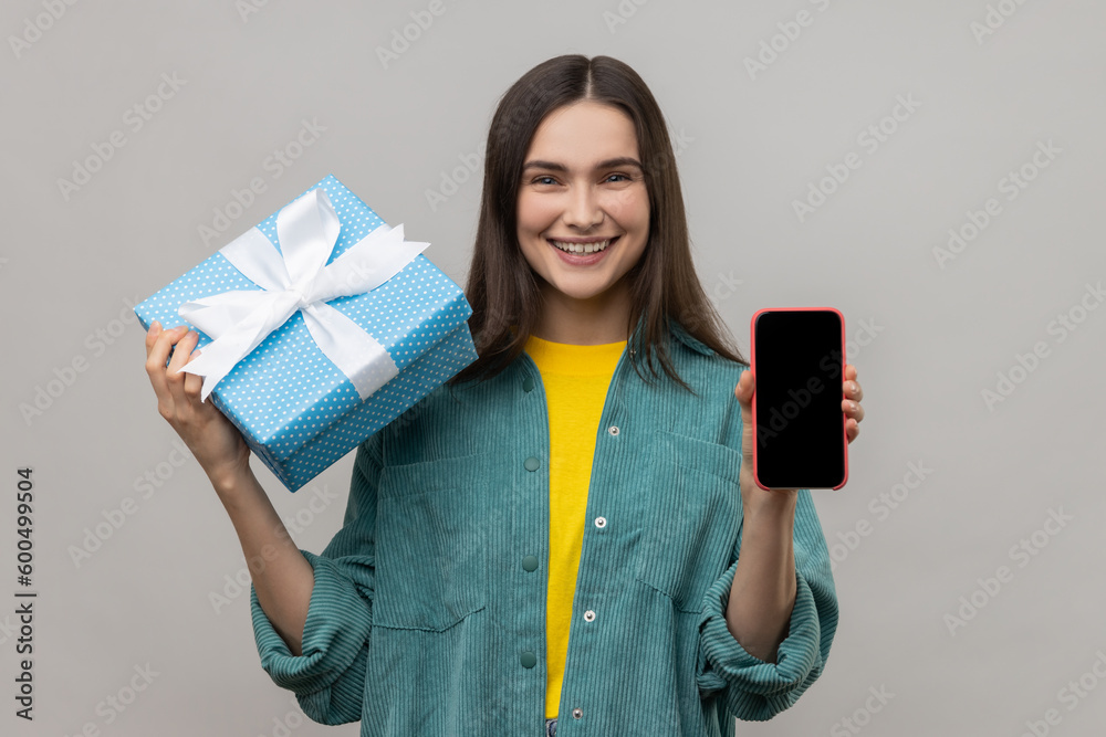 Present, bonus for mobile user. Woman holding gift box and cell phone with empty display for online shopping advertising, wearing casual style jacket. Indoor studio shot isolated on gray background. Present, bonus for mobile user. Woman holding gift box and cell phone with empty display for online shopping advertising, wearing casual style jacket. Indoor studio shot isolated on gray background.