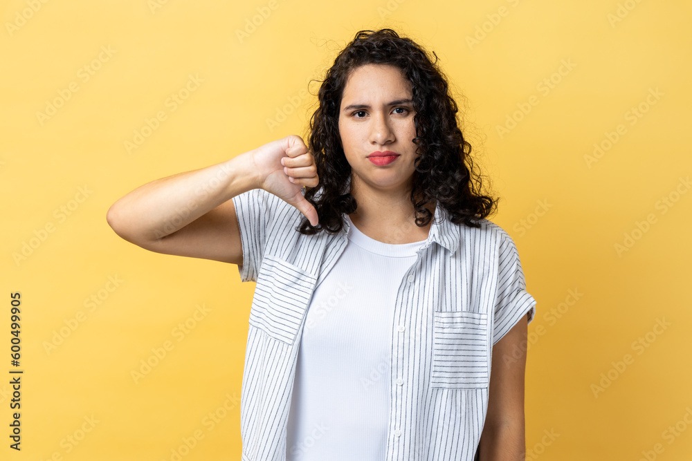 Portrait of woman with dark wavy hair criticizing bad quality with thumbs down displeased grimace, showing dislike gesture, expressing disapproval. Indoor studio shot isolated on yellow background. Portrait of woman with dark wavy hair criticizing bad quality with thumbs down displeased grimace, showing dislike gesture, expressing disapproval. Indoor studio shot isolated on yellow background.