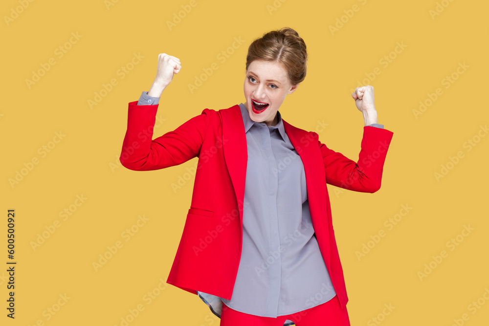 Portrait of extremely happy woman with red lips standing raised her clenched fists, screaming hurray, celebrating her victory, wearing red jacket. Indoor studio shot isolated on yellow background.