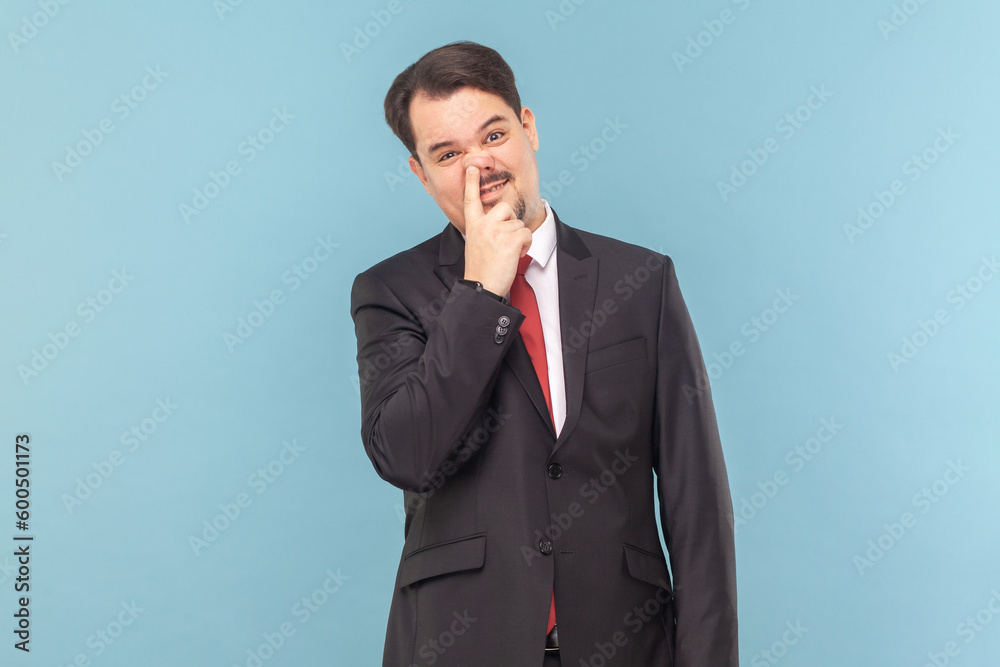 Portrait of funny childish man with mustache standing having comical facial expression, keeps finger in nose, wearing black suit with red tie. Indoor studio shot isolated on light blue background. Portrait of funny childish man with mustache standing having comical facial expression, keeps finger in nose, wearing black suit with red tie. Indoor studio shot isolated on light blue background.