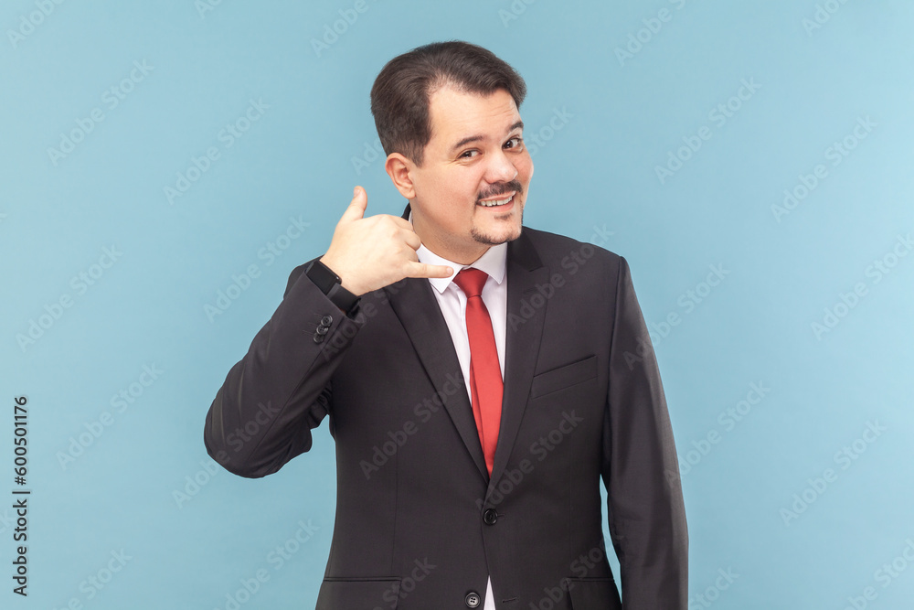Please, call me back. Portrait of optimistic cute smiling man with mustache standing and showing call me gesture, wearing black suit with red tie. Indoor studio shot isolated on light blue background. Please, call me back. Portrait of optimistic cute smiling man with mustache standing and showing call me gesture, wearing black suit with red tie. Indoor studio shot isolated on light blue background.