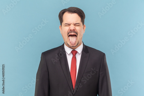 Portrait of crazy excited man with mustache standing sticking tongue out, demonstrates childish behavior, wearing black suit with red tie. Indoor studio shot isolated on light blue background.