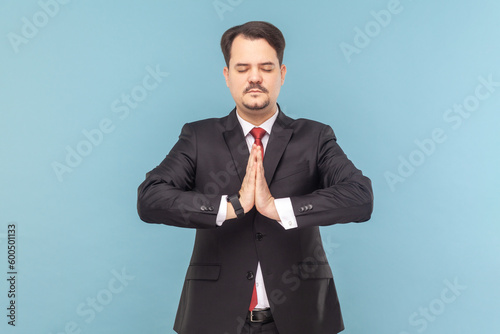 Portrait of calm man with mustache standing with closed practicing yoga, meditating while having break at work, wearing black suit with red tie. Indoor studio shot isolated on light blue background.