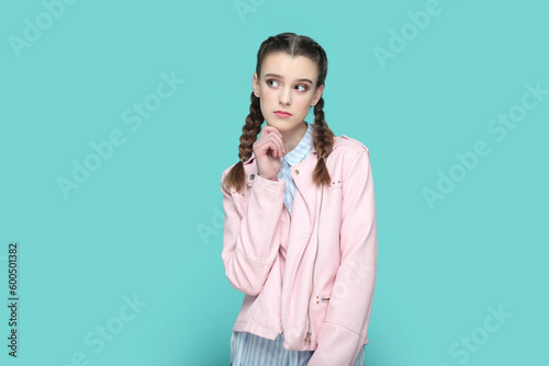 Portrait of pensive thoughtful teenager girl with braids wearing pink jacket standing holding her chin, looking away, planning her future. Indoor studio shot isolated on green background.