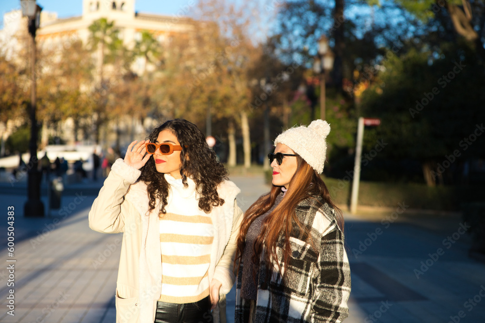 two young, beautiful, Spanish women who are friends dressed in winter clothes and wearing sunglasses are walking and enjoying the sunny day in seville, spain.